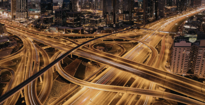 Aerial night view of a large multi-level urban highway interchange with glowing light trails from traffic and an illuminated city skyline in the background