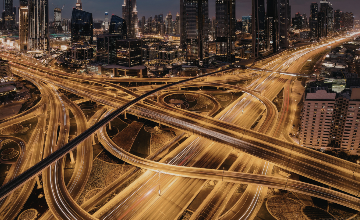 Aerial night view of a large multi-level urban highway interchange with glowing light trails from traffic and an illuminated city skyline in the background