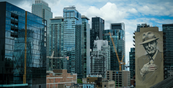 Downtown Montreal skyline showing glass high-rise towers, active construction cranes, and the large Leonard Cohen mural on a building facade under a cloudy sky