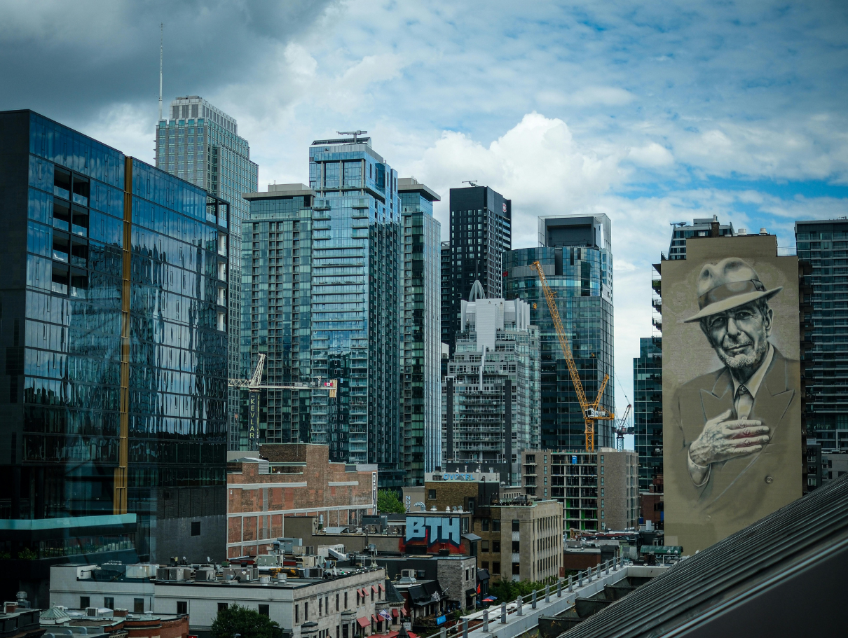 Downtown Montreal skyline showing glass high-rise towers, active construction cranes, and the large Leonard Cohen mural on a building facade under a cloudy sky