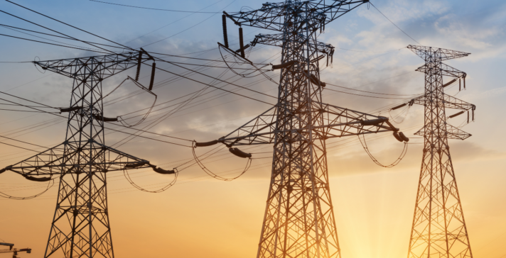 Silhouetted high-voltage electrical transmission towers and power lines against a glowing orange and blue sunset sky