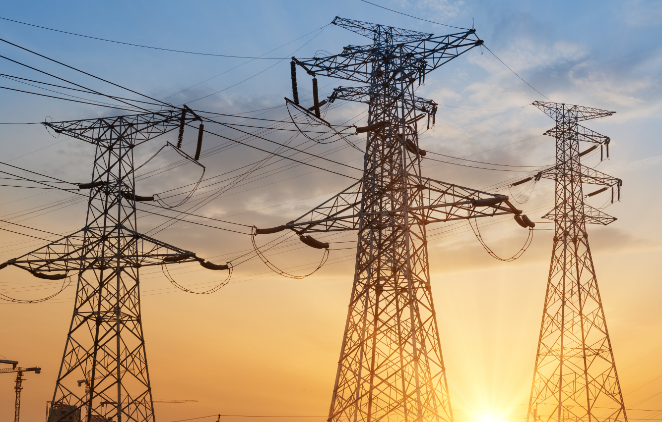 Silhouetted high-voltage electrical transmission towers and power lines against a glowing orange and blue sunset sky
