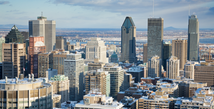 Aerial view of the downtown Montreal skyline in winter, with snow-dusted rooftops, high-rise towers, and the St. Lawrence River in the distance