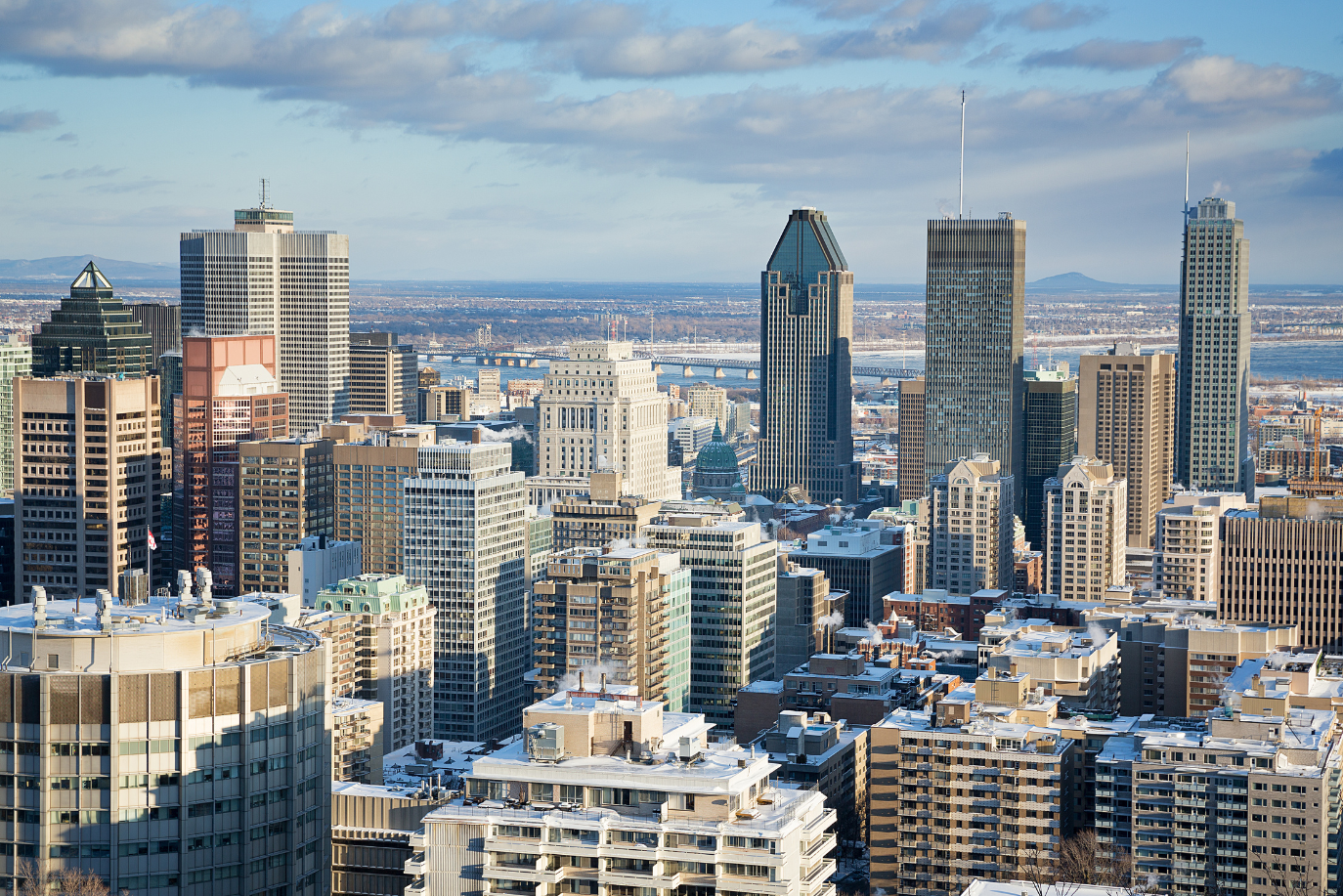 Aerial view of the downtown Montreal skyline in winter, with snow-dusted rooftops, high-rise towers, and the St. Lawrence River in the distance
