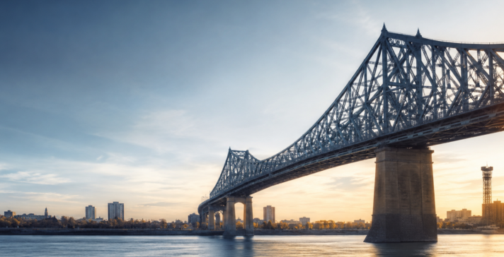 Realistic view of a large steel bridge crossing a calm river at dawn, with soft blue and golden light and a distant city skyline.