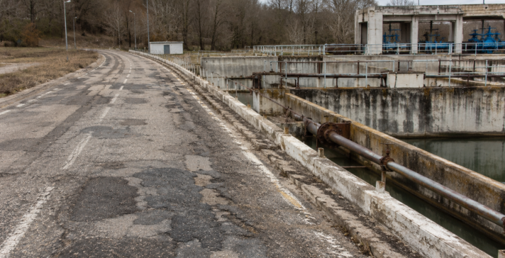 Weathered roadway with cracked and patched asphalt beside an aging water treatment structure with stained concrete, rusted pipes, railings, and water channels.