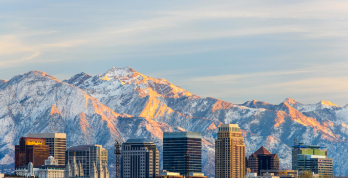 Downtown city skyline of high-rise buildings backed by snow-capped mountains glowing in warm light under a soft blue sky
