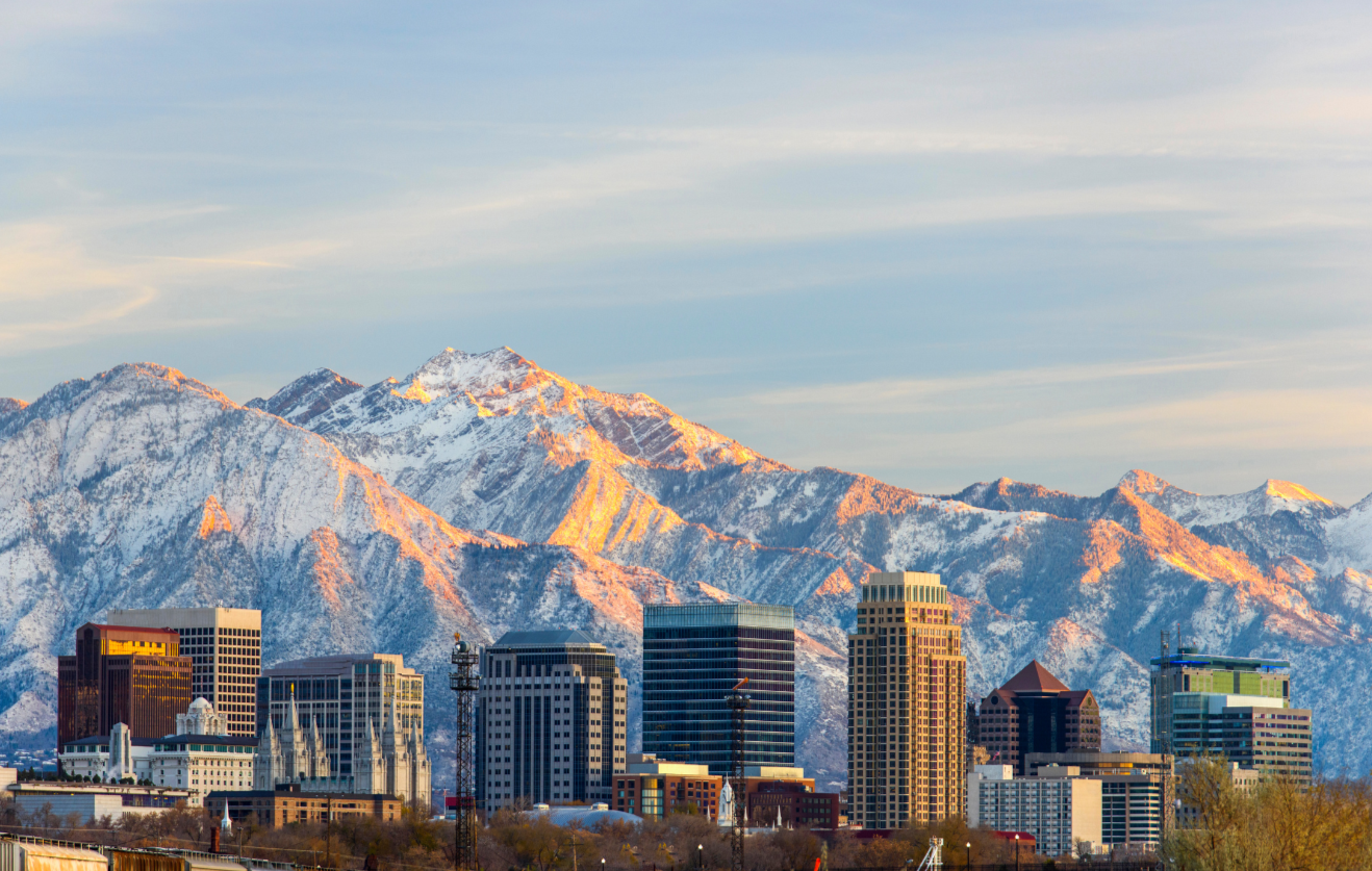 Downtown city skyline of high-rise buildings backed by snow-capped mountains glowing in warm light under a soft blue sky