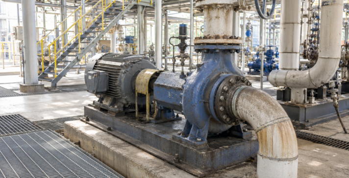 Bright industrial facility showing an aged blue pump, motor, pipes, valves, metal grating, and yellow safety railings.