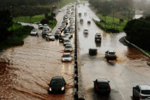 Cars drive through a flooded road wherein a city's water infrastructure has been damaged due to climate change.