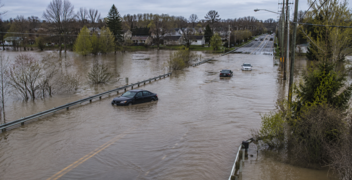 Flooded road outside a city with partially submerged cars, guardrails, utility poles, trees, and nearby houses under an overcast sky.