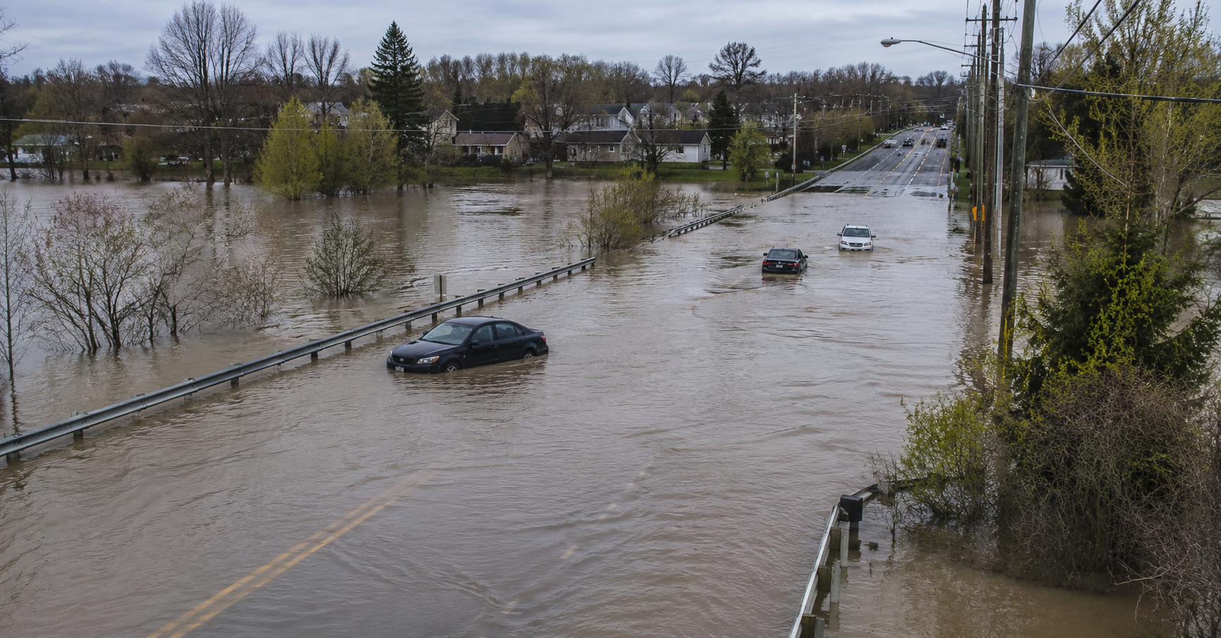 Flooded road outside a city with partially submerged cars, guardrails, utility poles, trees, and nearby houses under an overcast sky.