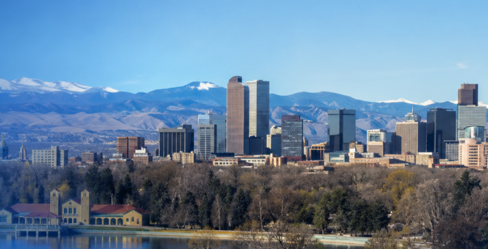 Wide view of the Denver skyline with mountains in the background, a lake and park in the foreground, and a blue brand-style overlay.