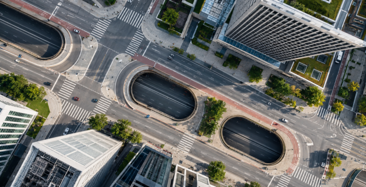 Directly overhead aerial view of a city intersection with roads, crosswalks, oval tunnel entrances, and surrounding high-rise buildings