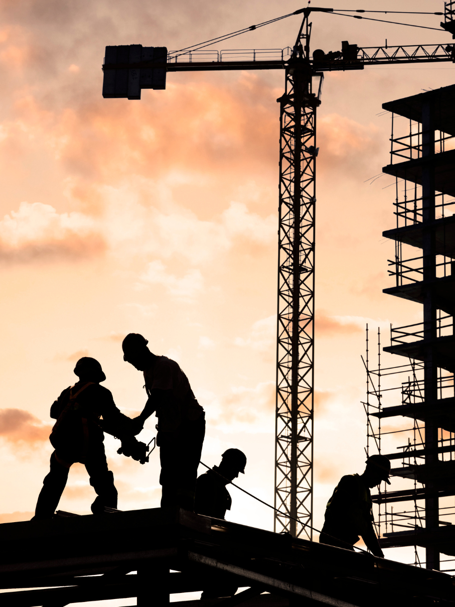 Silhouettes of construction workers on a high-rise building site with a tower crane against a sunset skyConstruction workers at height on a high-rise site, framed against a tower crane and sunset, representing the scale and coordination behind municipal infrastructure projects.