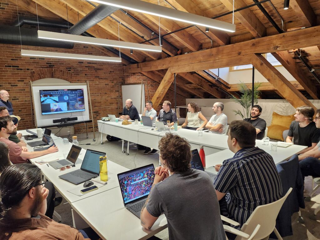 Team gathered around conference tables during a collaborative meeting in a modern loft-style office with exposed wood beams and brick walls.