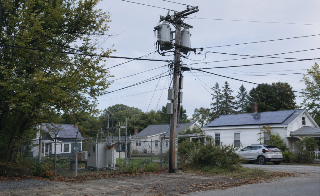 Wooden utility pole with two pole-mounted distribution transformers and overhead lines in front of homes with rooftop solar panels