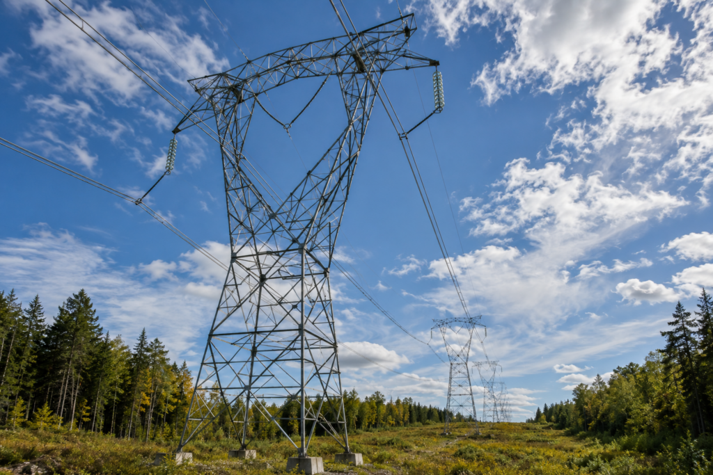 Lattice steel transmission towers carrying high-voltage lines along a cleared corridor between forested edges