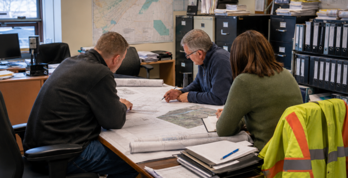 Three municipal staff reviewing engineering drawings and a site map during an infrastructure asset management planning meeting