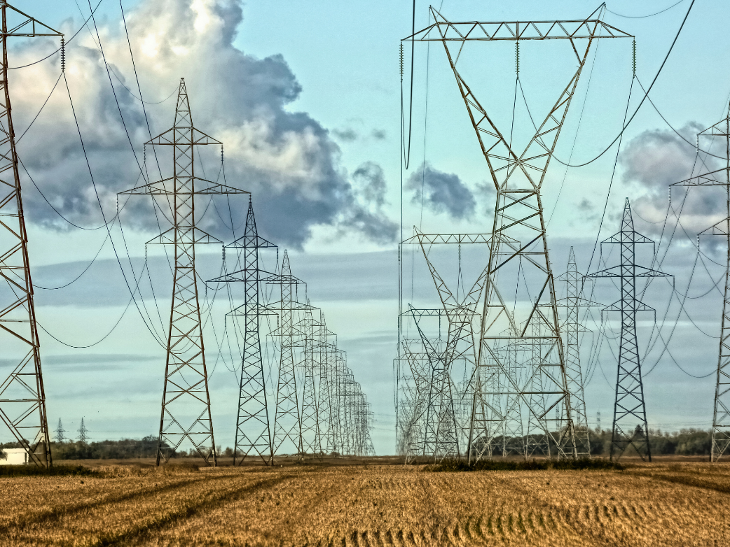 Multiple parallel rows of steel transmission towers carrying high-voltage lines across a harvested field