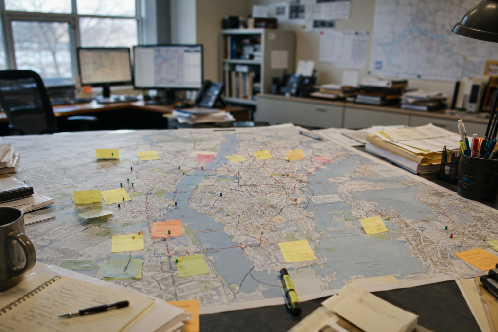 Large city map spread across a desk, marked with colored sticky notes and pushpins in a planning office