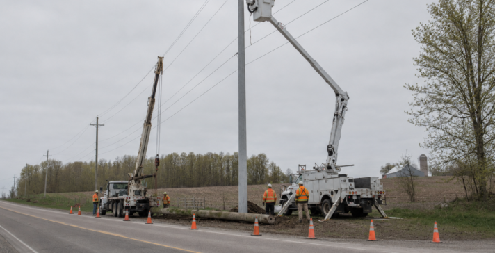 Utility crew replacing a wooden distribution pole with a taller reinforced pole along a rural road, old pole on the ground