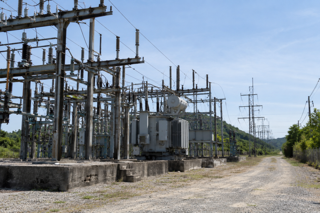 Daytime view of an aging electrical substation with transformers, steel structures, overhead power lines, and a gravel access road.