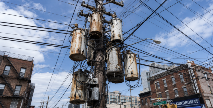 Aging utility pole with weathered distribution transformers and overhead power lines above an older city street.