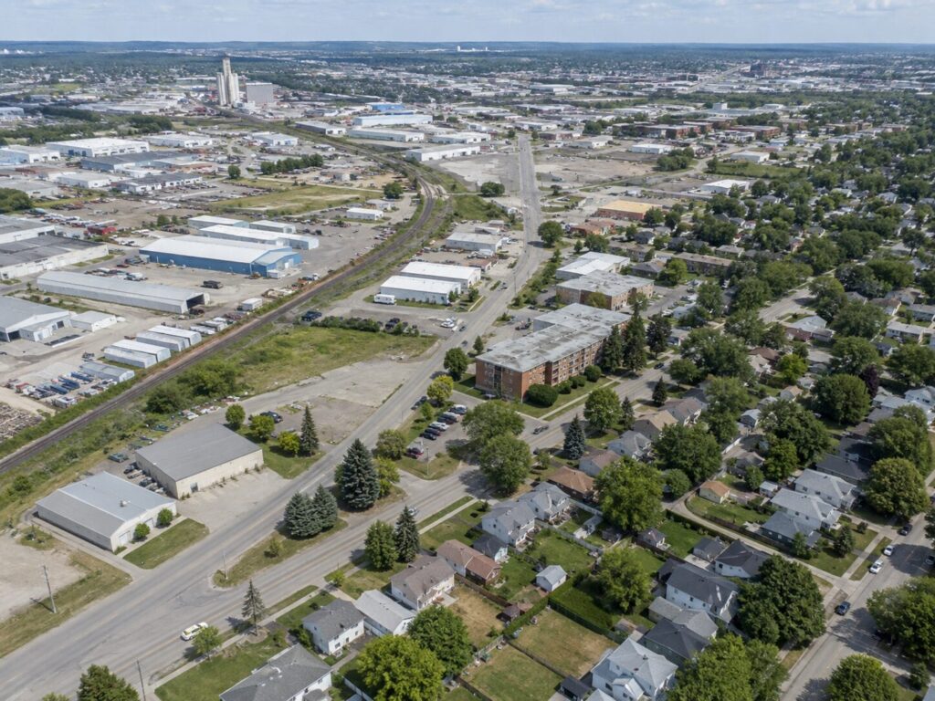 Aerial photograph showing an industrial park with warehouses and rail lines on the left transitioning into tree-lined residential streets with single-family homes on the right