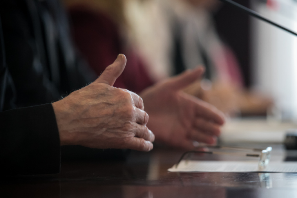 Close-up of hands gesturing at a council meeting table with a microphone and documents, representing an infrastructure budget discussion