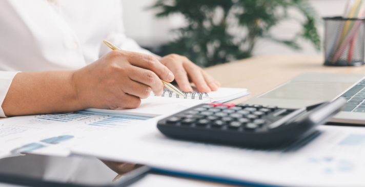 Person writing in a notebook at a desk with a calculator, laptop and printed financial charts, reviewing an infrastructure budget