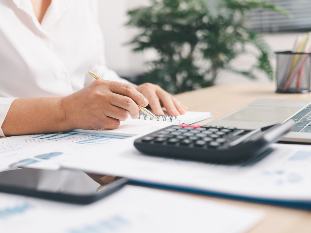 Person writing in a notebook at a desk with a calculator, laptop and printed financial charts, reviewing an infrastructure budget