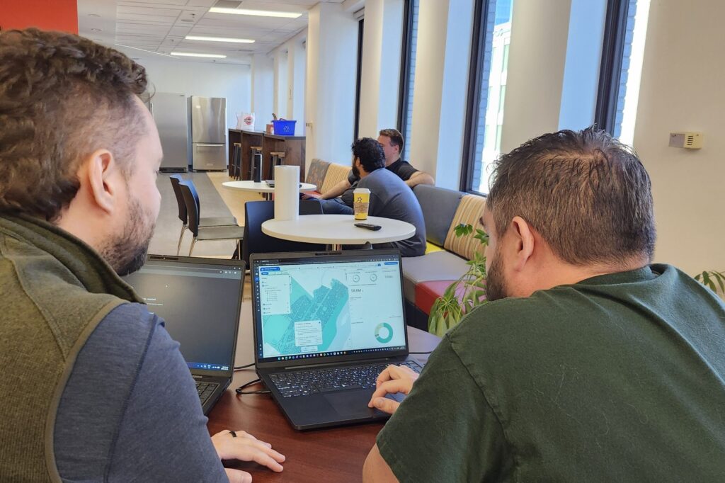 Two colleagues at a table reviewing a map-based asset planning dashboard on a laptop in a bright open office.